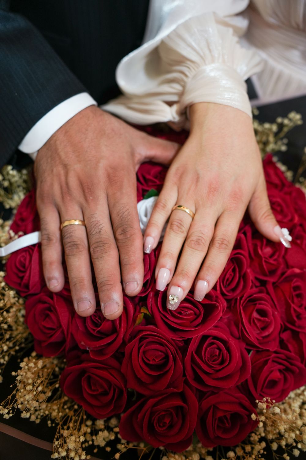 Fotografía de muestra: manos con anillos de boda sobre un ramo de rosas rojas.